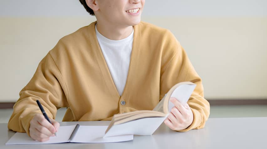机に向かってノートにペンを走らせながら、笑顔で顔を上げるマスタード色のカーディガンを着た若い男性。手元には開いた本があり、前向きに学習に取り組んでいる様子や、何かを発見して喜んでいるシーンを表現している。