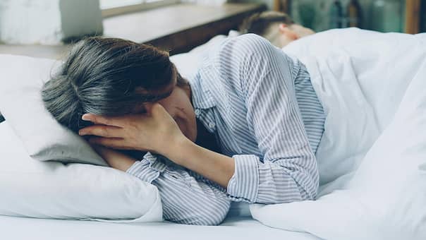ベッドに横たわり、両手で顔を覆って悩んでいるような様子の女性。ストライプのパジャマを着用しており、不眠、頭痛、あるいは精神的なストレスを感じている重苦しい雰囲気を伝えている。