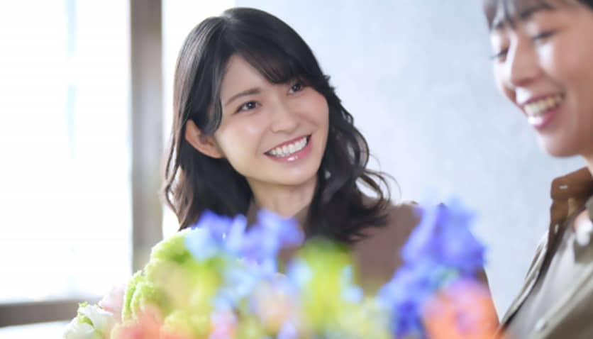 屋内で花を前にして楽しそうに談笑する二人の女性。手前の女性は明るい笑顔で相手を見つめており、ポジティブな対人関係や、おしゃべりを楽しむ和やかな雰囲気を伝えている。