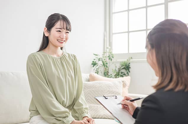 明るい室内でソファに座り、笑顔で対話する若い女性。手前にはバインダーにメモを取る担当者の後ろ姿があり、カウンセリングやキャリア相談、インタビューを行っている穏やかなシーンを表現している。