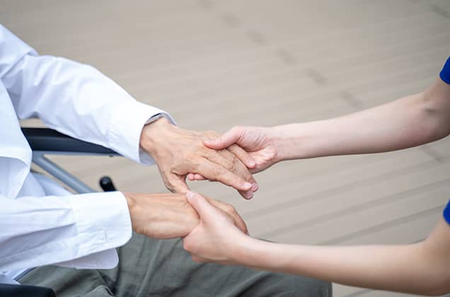 車椅子に座る高齢と思われる方の両手を、介助者の手が優しく包み込むように握っているクローズアップ写真。心のこもった介護、看護、あるいは家族の絆と安心感を象徴している。